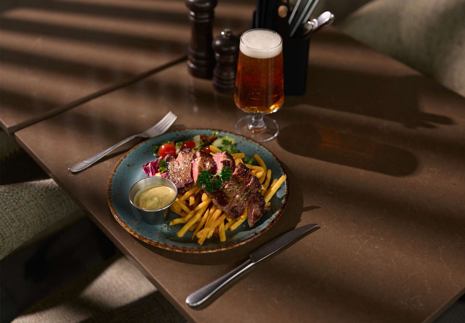 Plate with sliced steak, fries, salad and a small bowl of sauce, served with a glass of beer.