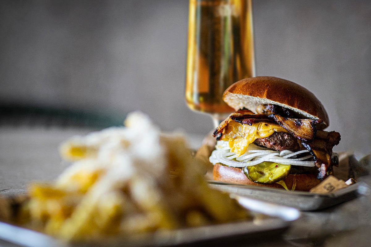 Burger with cheese and bacon on a plate, with fries in the foreground and a glass of beer behind.